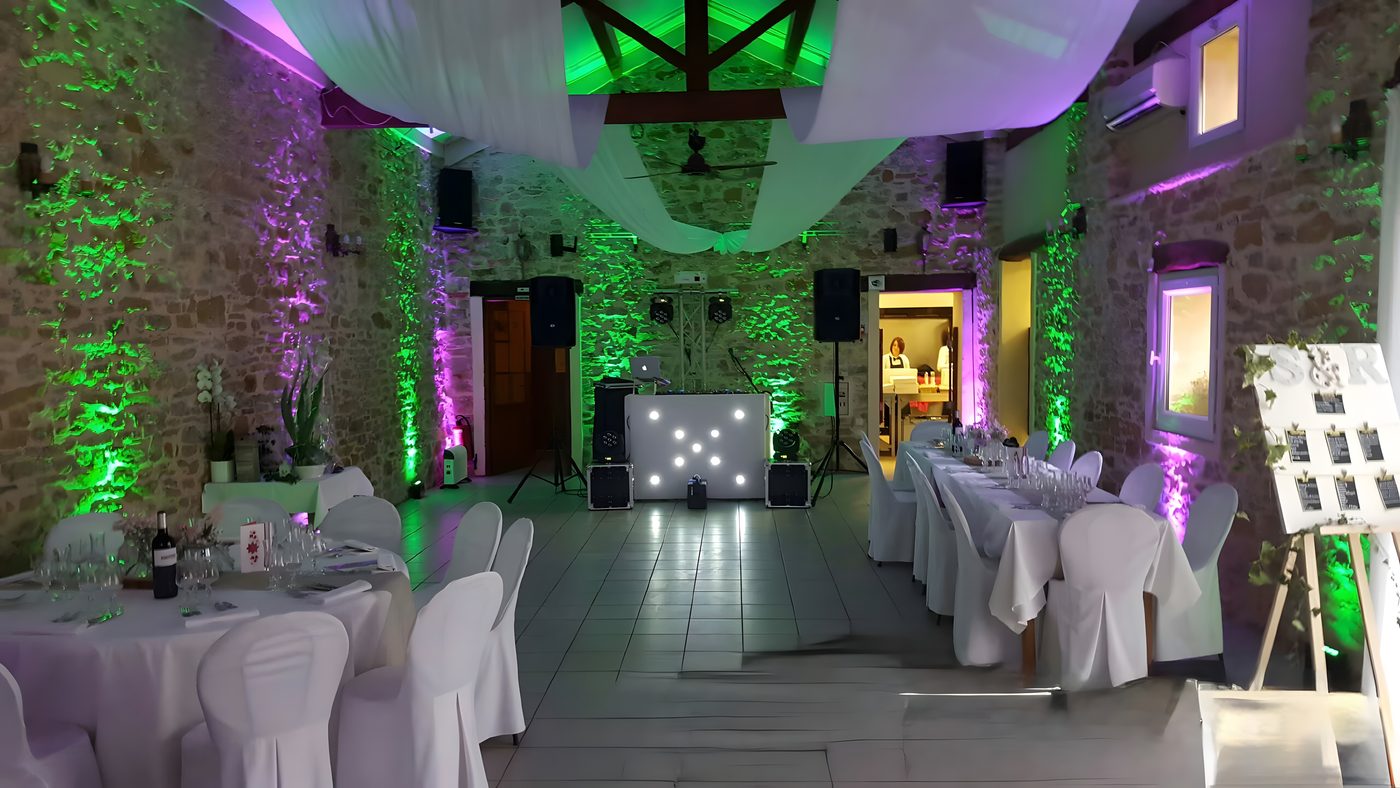 Salle de réception sous charpente, drapé blanc au plafond, murs de pierre éclairés en vert et violet, régie du DJ au fond et tables dressées en blanc