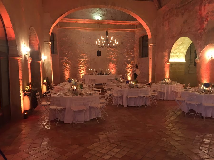 Grande salle voûtée en pierre d’un domaine, lustre en fer forgé, murs éclairés en ambre et tables rondes dressées avant le repas