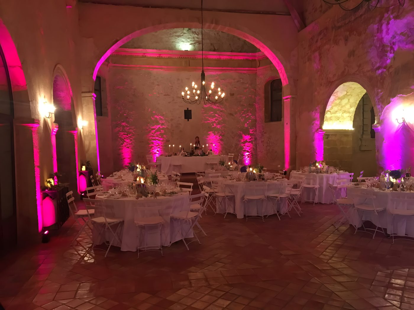 La même salle voûtée en pierre, éclairée cette fois en rose et magenta, lustre allumé et tables rondes dressées