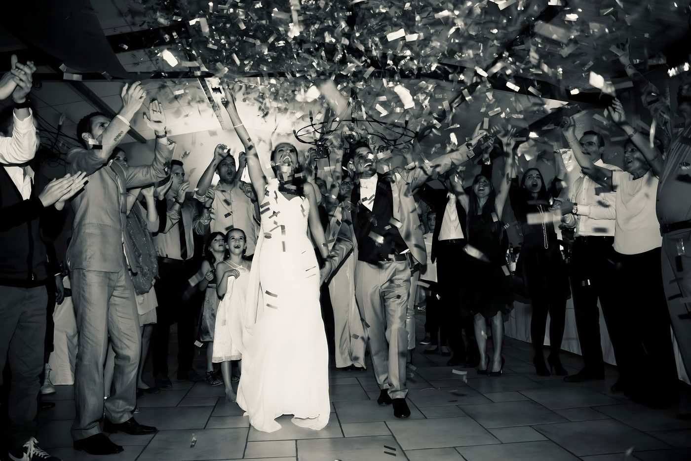 Pluie de confettis sur les mariés au milieu des invités bras levés, en noir et blanc