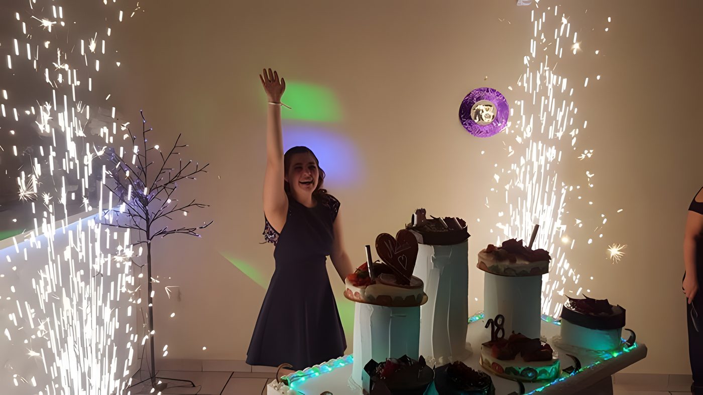 Table de desserts d’anniversaire entre deux fontaines d’étincelles, boule à facettes violette et arbre lumineux, la personne fêtée lève le bras