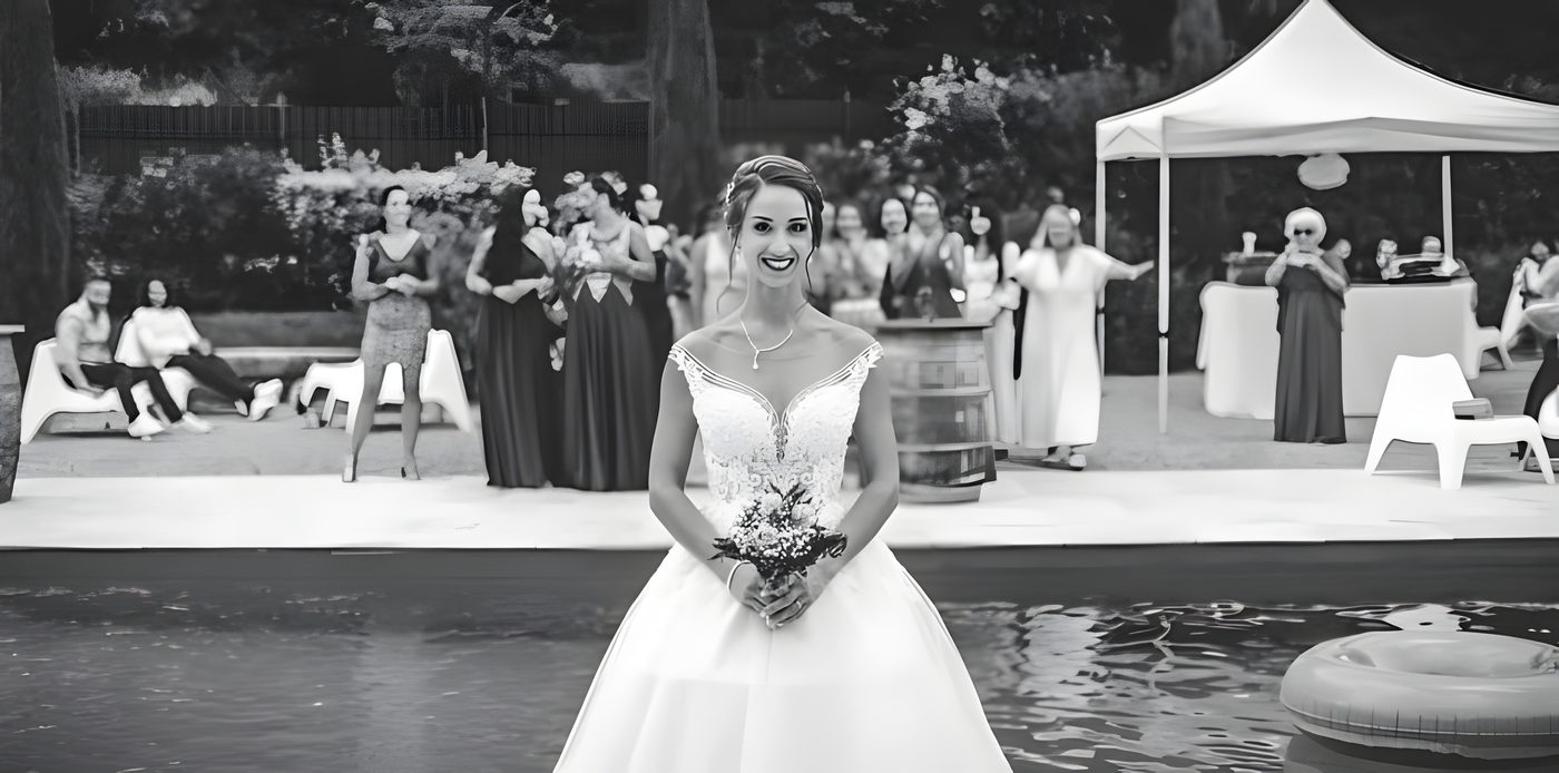 La mariée debout au bord d’une piscine, bouquet à la main, invités en arc de cercle et barnum blanc sous les arbres, en noir et blanc