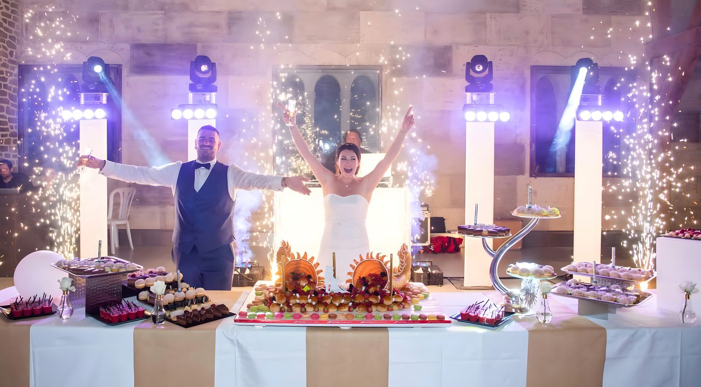 Les mariés bras ouverts derrière la longue table des desserts, totems lumineux, lyres sur pied et jets d’étincelles froides