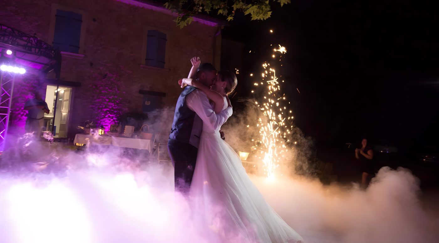 Baiser des mariés en extérieur devant une bastide en pierre, fumée lourde teintée de rose et jet d’étincelles froides sur la droite