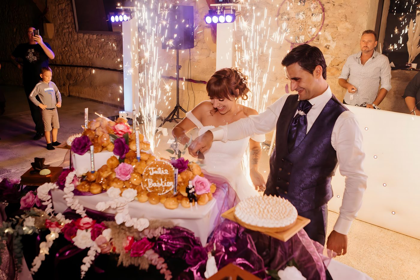 Les mariés découpent une pièce montée de choux sur une table fleurie, jet d’étincelles froides derrière eux et invités autour