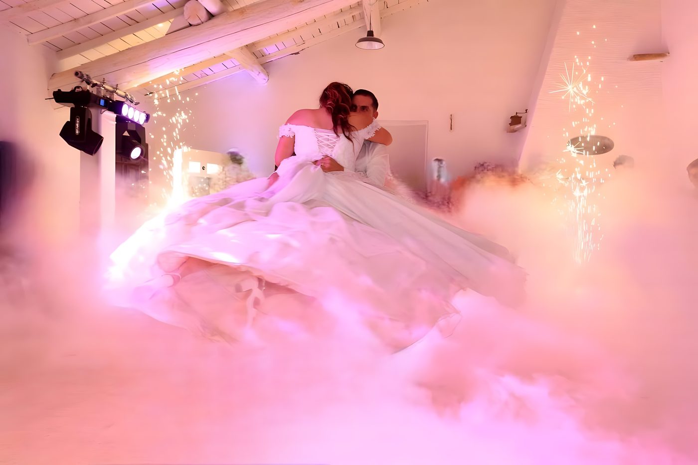 La mariée dans les bras du marié, sa robe étalée sur la fumée lourde rose, deux jets d’étincelles froides dans une salle blanche sous charpente